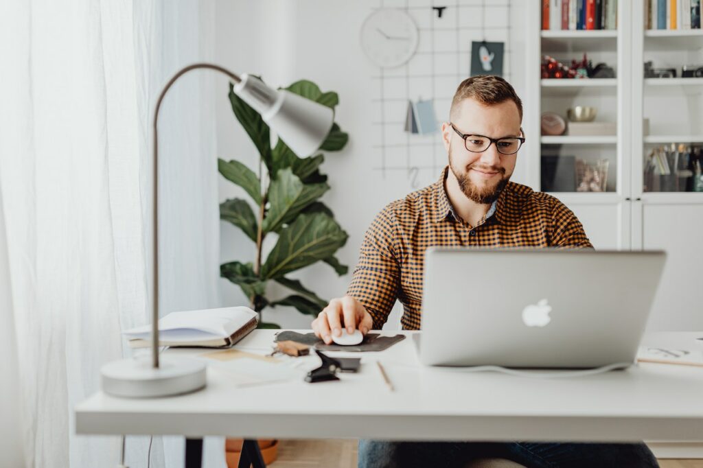 A man in a yellow and blue checkered polo shirt using a silver MacBook and a white mouse with a lamp and office supplies on a white table in the office