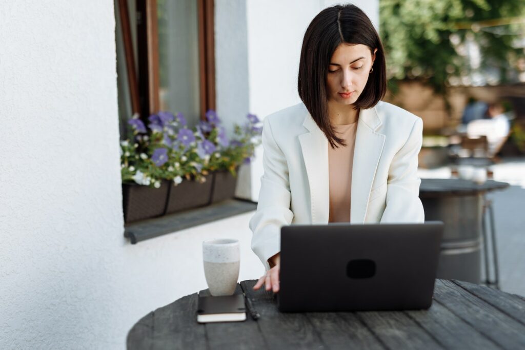 A woman using a gray laptop beside a black notebook and a white mug on a black wooden table outside the house