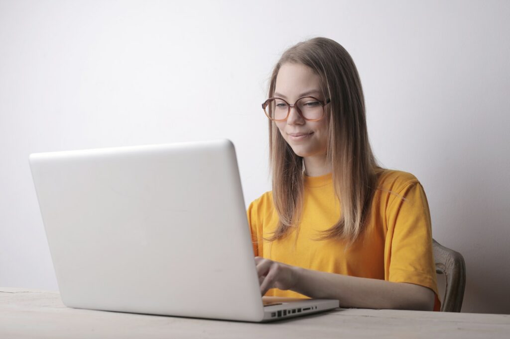 A white laptop being used by a woman wearing a yellow crew-neck t-shirt sitting on a wooden chair