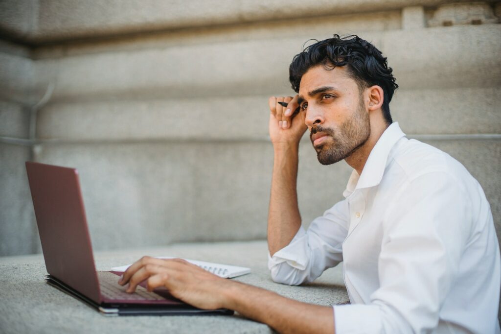 A man in white long-sleeves using a pink laptop and notebook placed on a cemented table