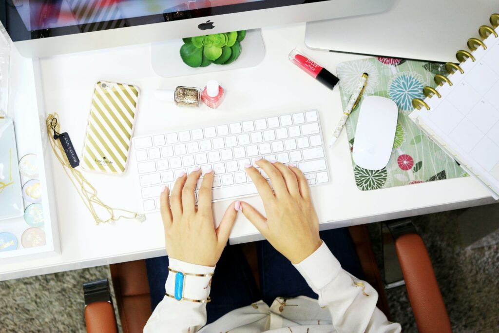 A woman with pink nail polish is using her iMac near a white smartphone, a gold necklace, and pink and glittery gold nail polish on top of a white wooden table