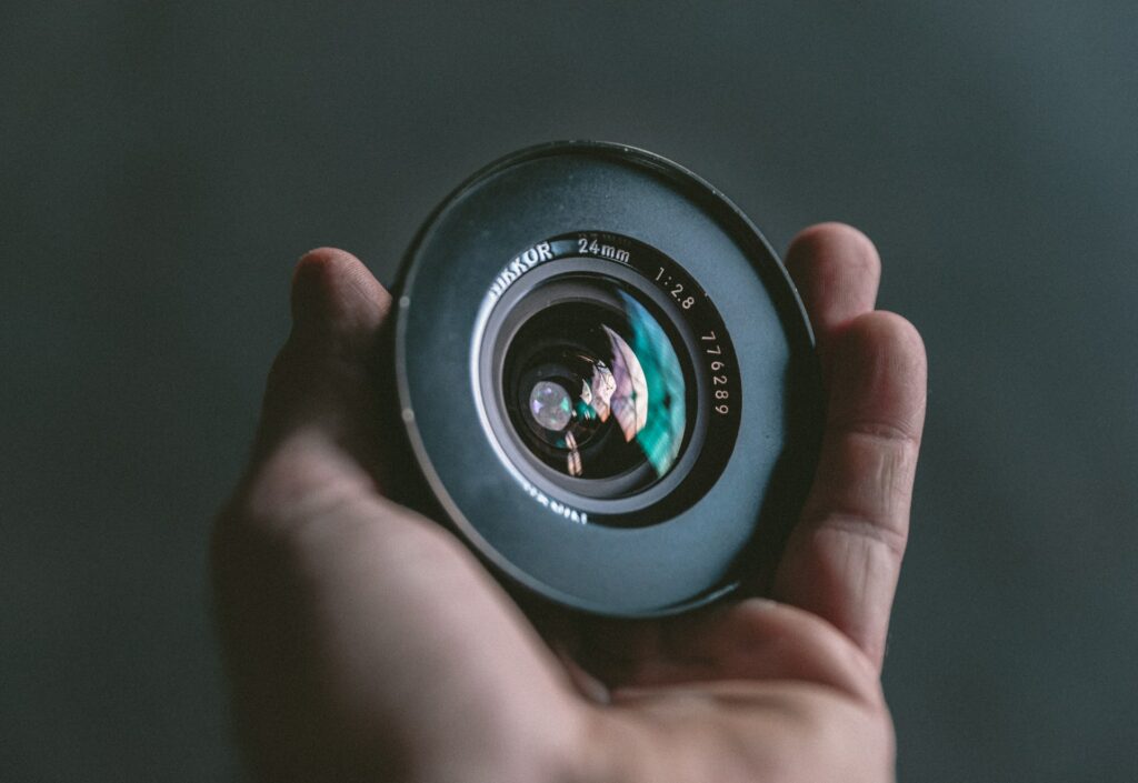 A person holding a black Nikkor 24mm camera lens near a black wall