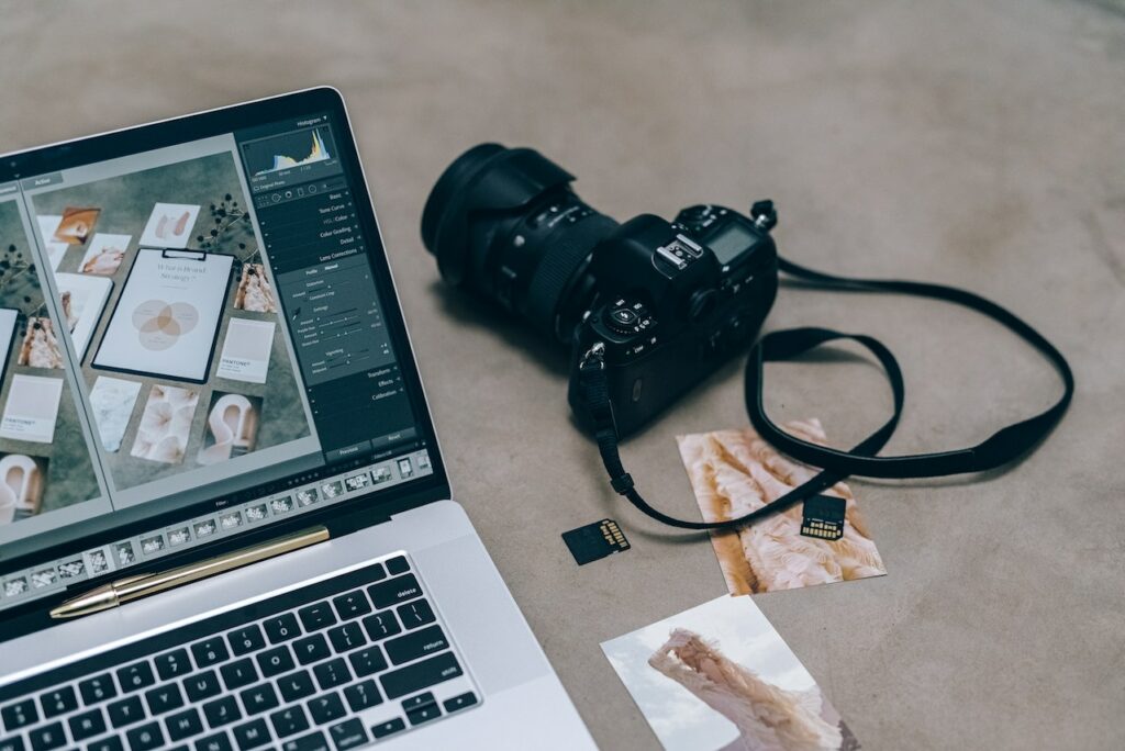 A black DSLR camera and silver laptop beside memory cards and pictures on a brown surface