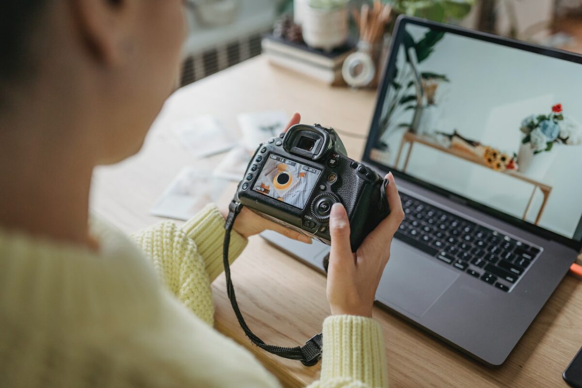 A person wearing a yellow sweatshirt is holding a black DSLR camera near a silver laptop that is placed on top of a brown wooden table