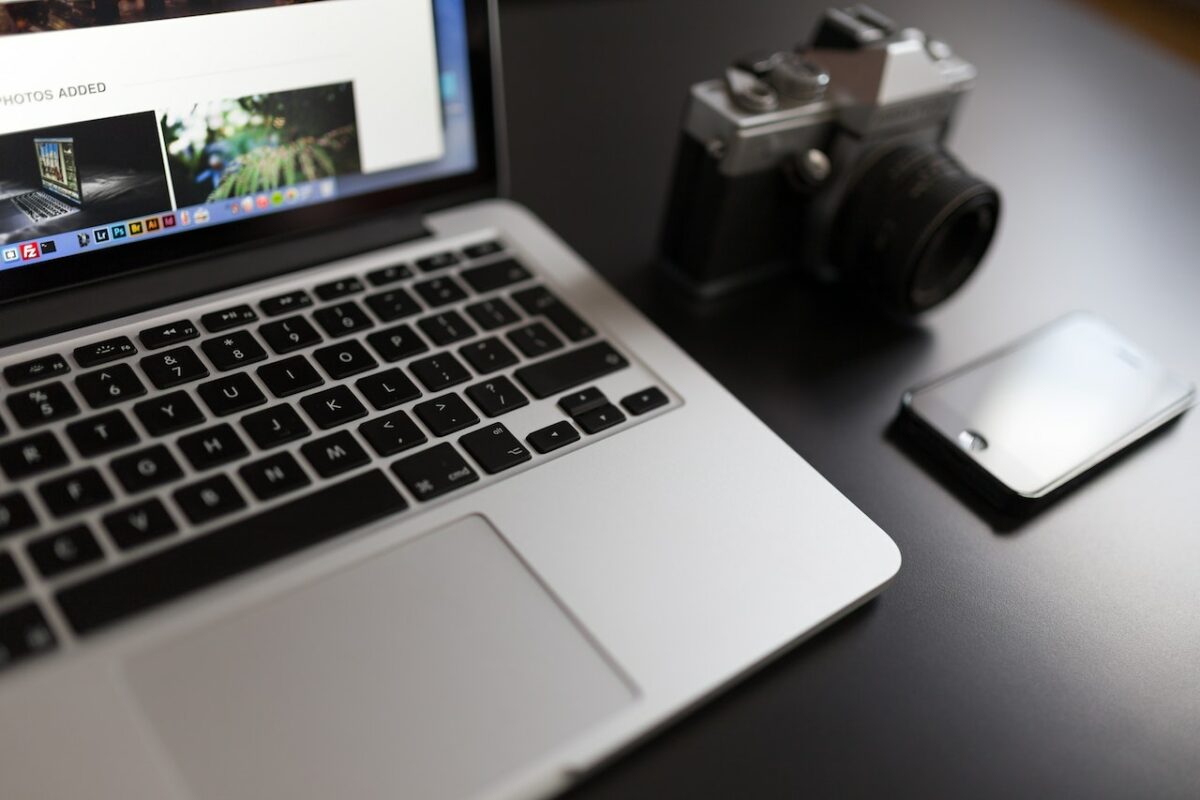 A silver laptop near a black and silver camera and smartphone on top of a black table