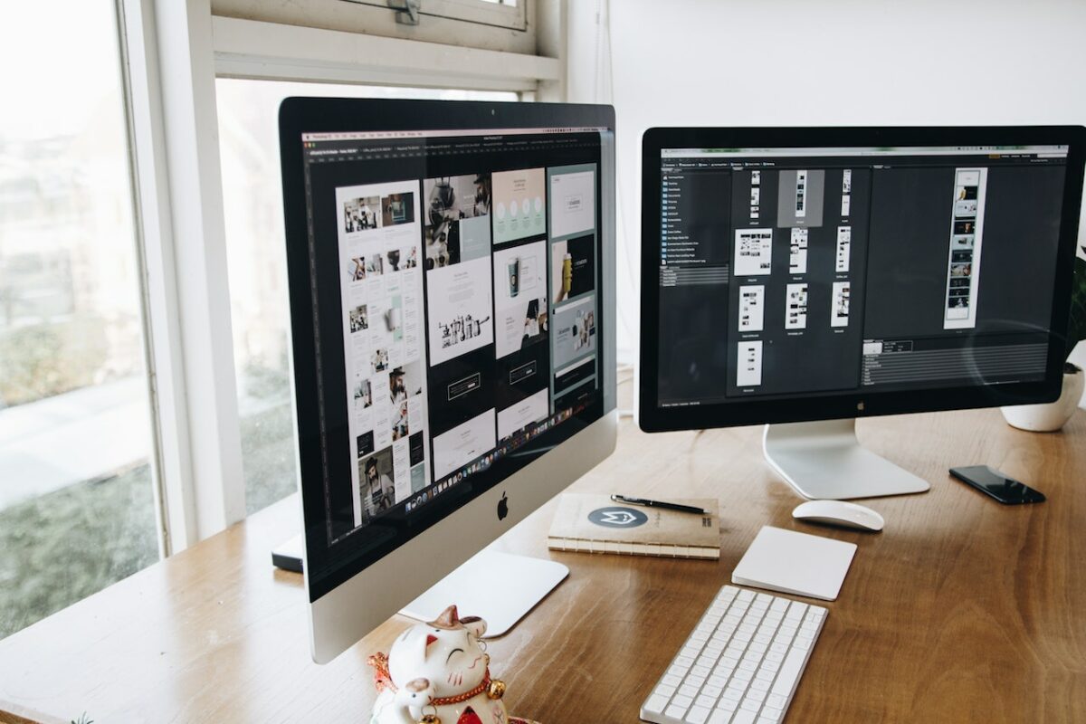 Zilveren en zwarte iMacs bij een wit toetsenbord en muis bovenop een bruine houten tafel bij het raam