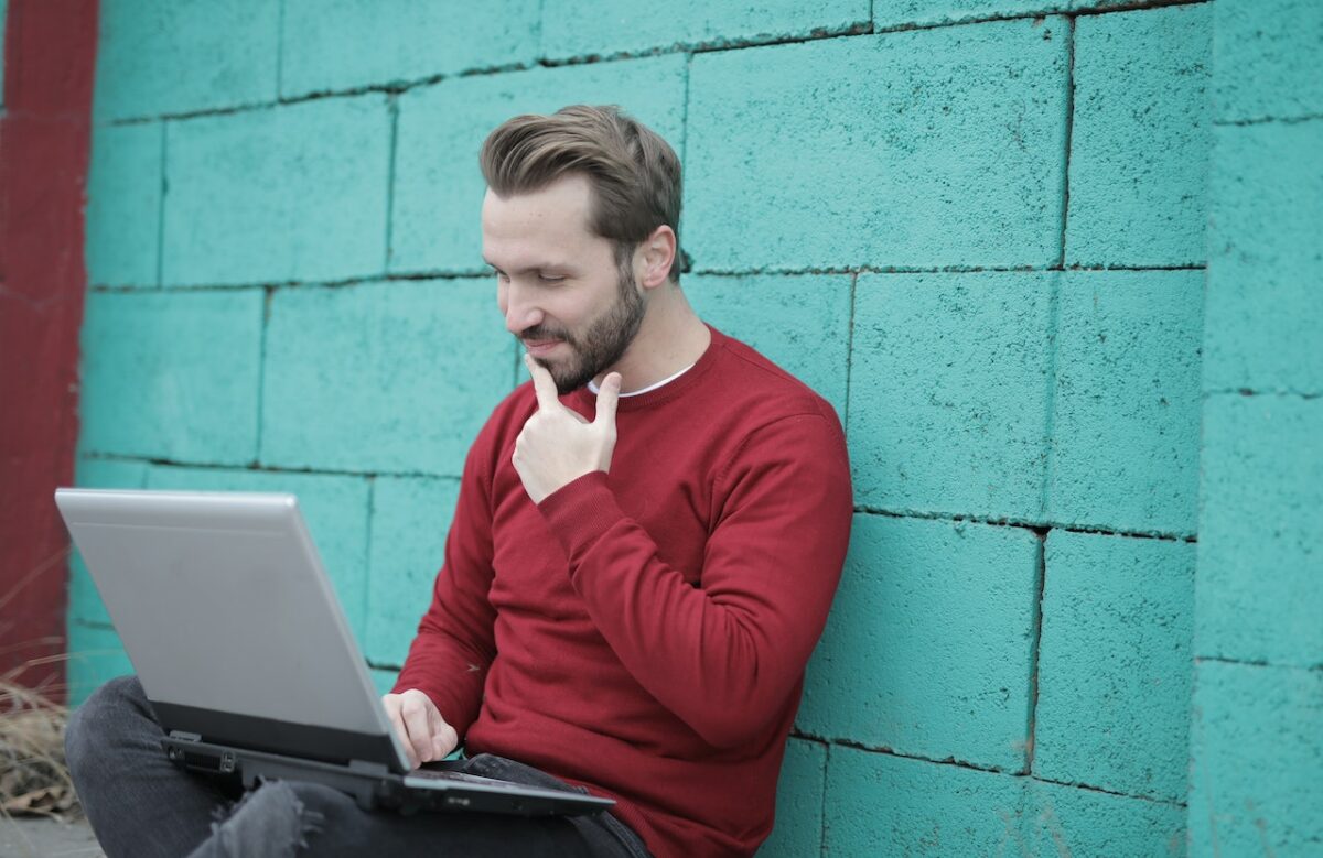 A man in a red long-sleeves and black pants uses a gray laptop while seated on a cemented floor