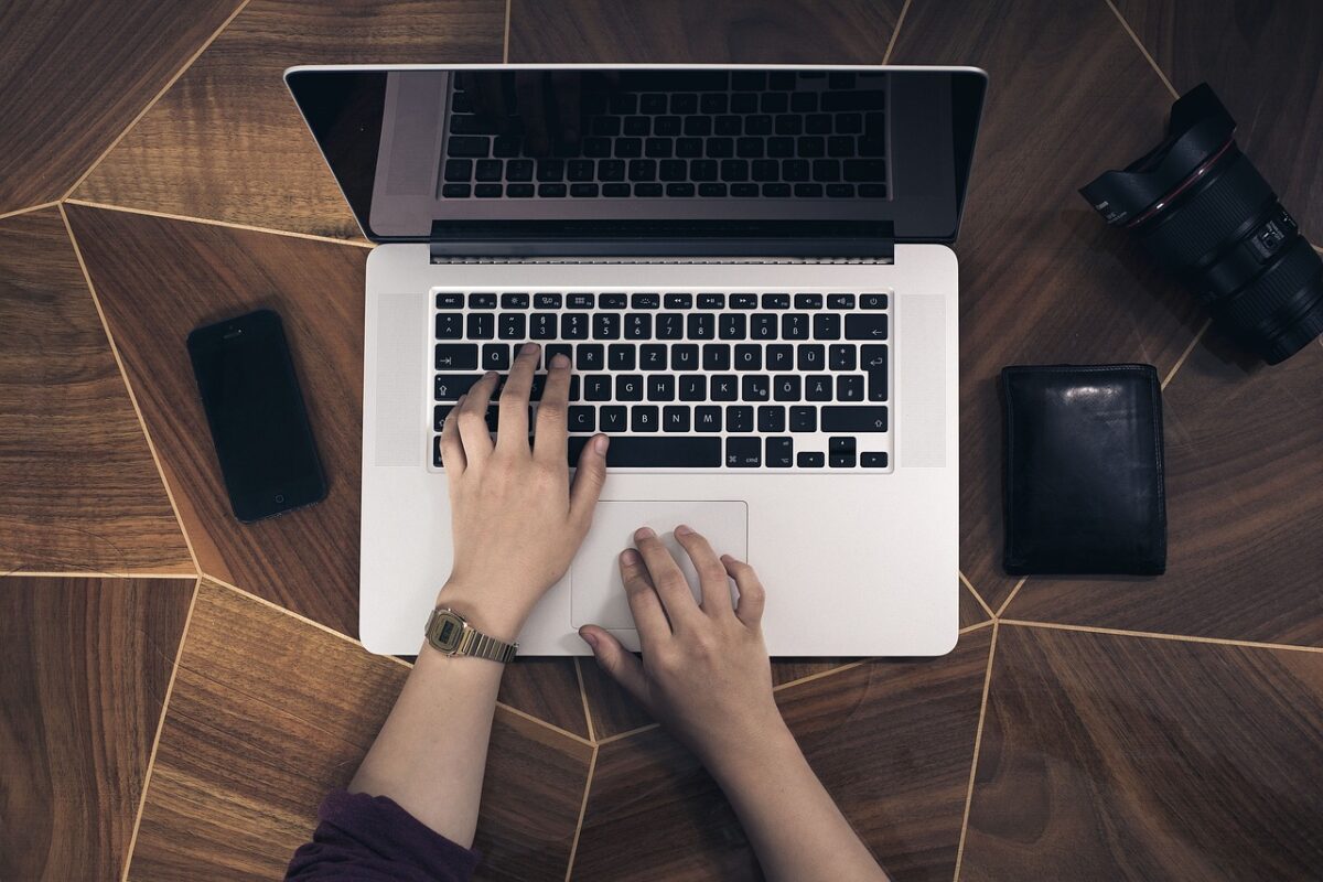 A person using a silver laptop near a black phone, black wallet, and DSLR camera on top of a wooden table