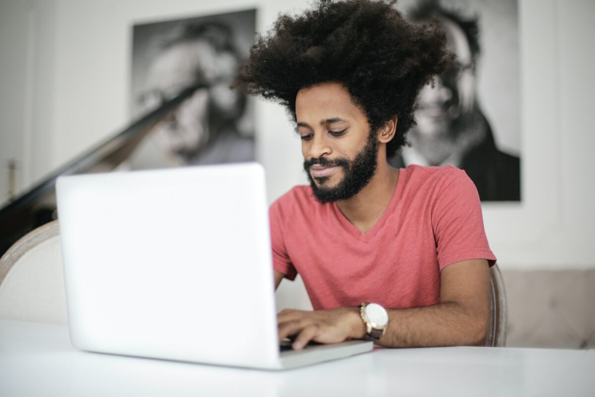 A man in a pink crew neck t-shirt uses a silver laptop on top of a white table