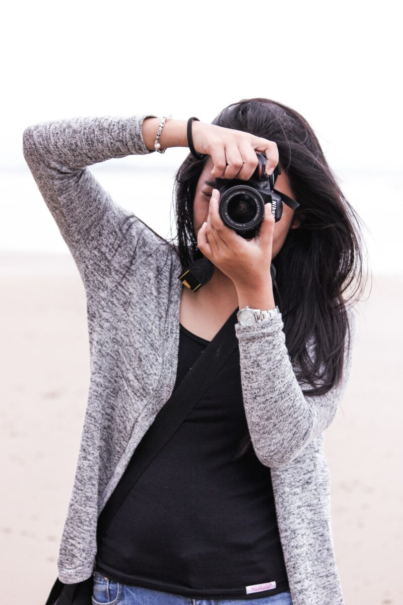 A woman in a gray cardigan and black shirt using a black Nikon DSLR camera on capturing photos