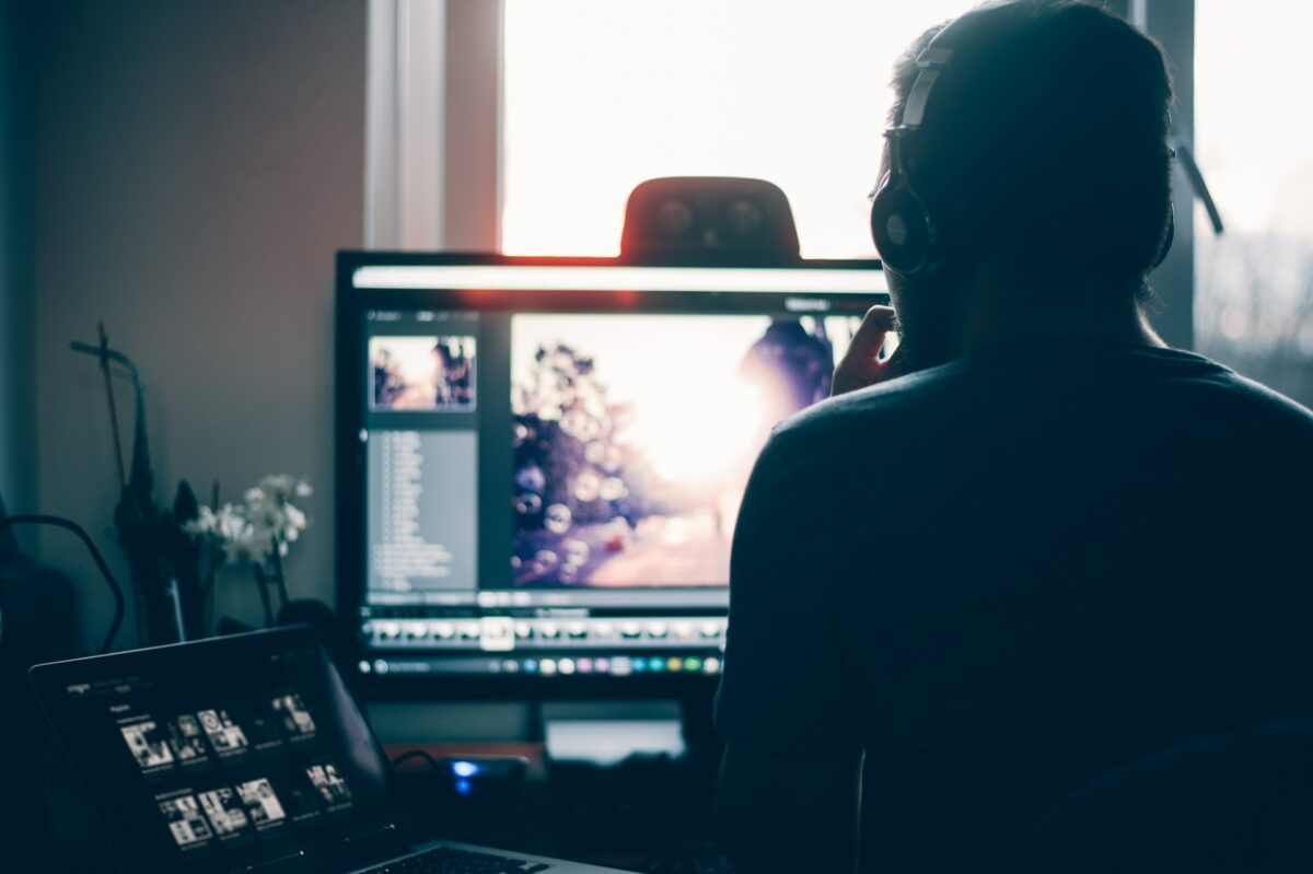 A man wearing silver headphones is using a silver laptop and black monitor placed near a window