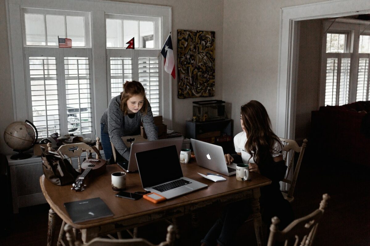 A woman wearing gray long sleeves shirt is working together with a woman wearing a white shirt while using a silver MacBook placed on top of a brown wooden table