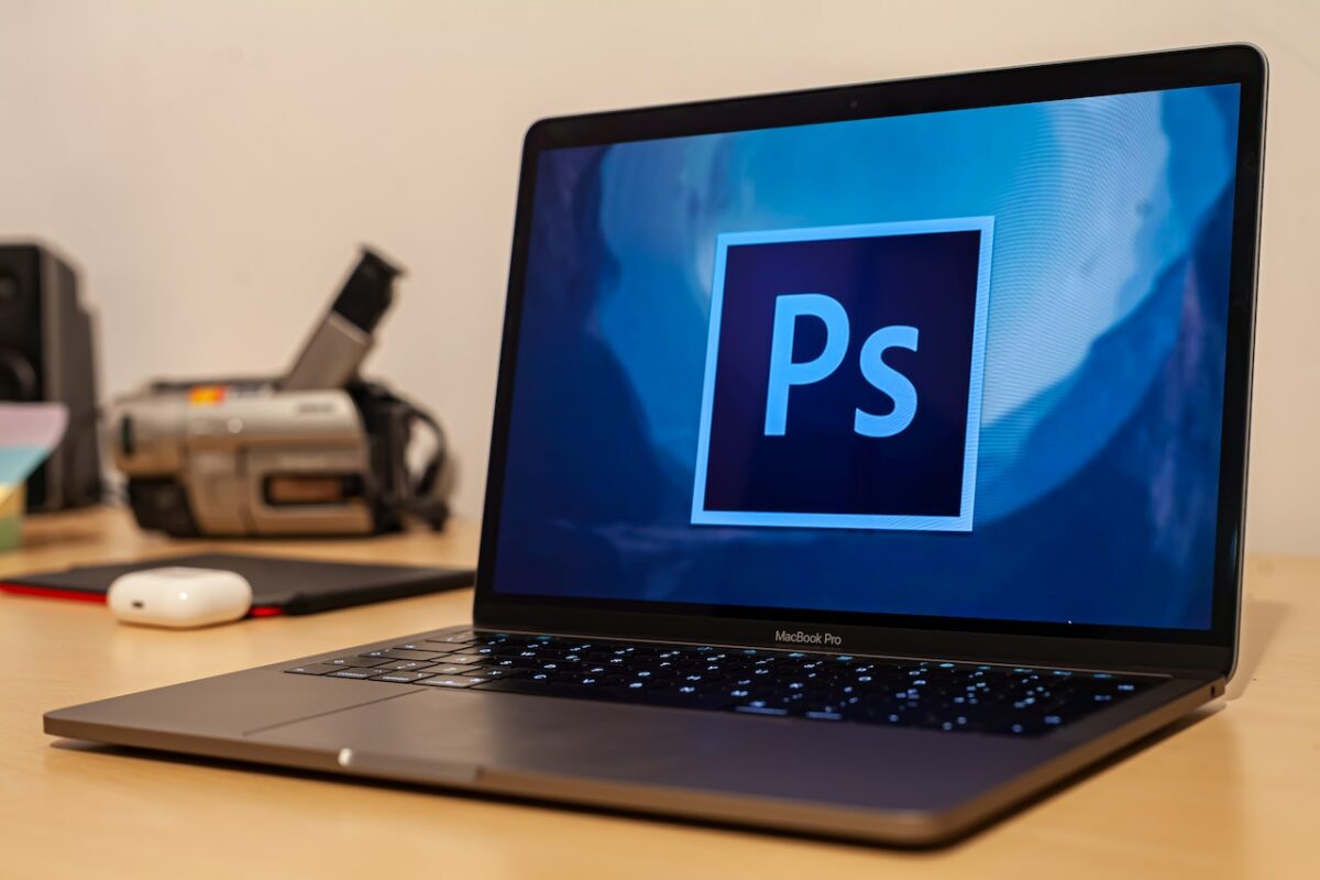 A silver MacBook Pro with the Adobe Photoshop logo on the screen on top of a brown wooden table near a white wall