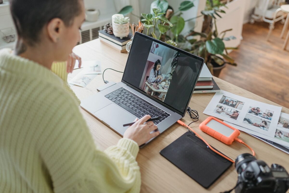 A woman in yellow sweatshirt editing pictures on a silver laptop placed on a brown wooden table