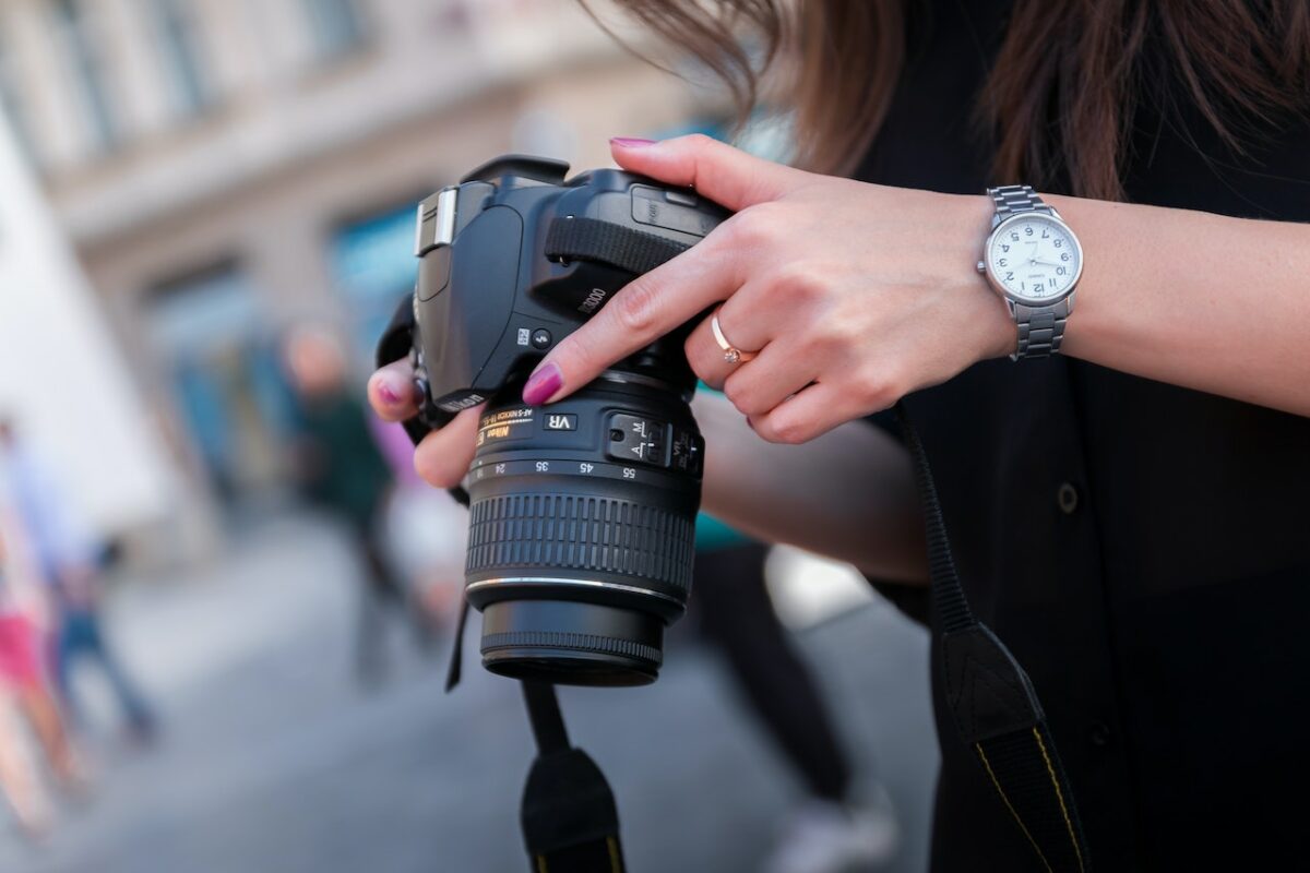 A woman wearing a black shirt and silver watch is holding a black DSLR camera in a public place