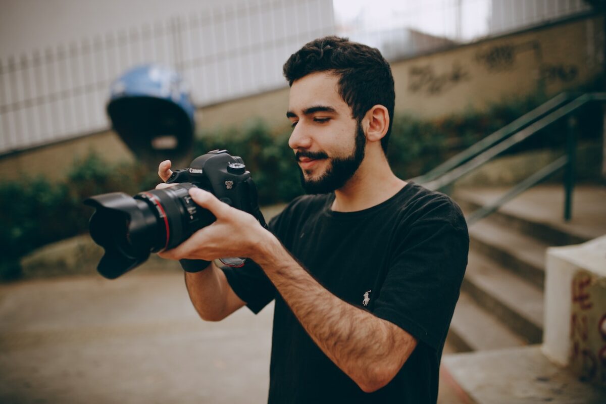 A man wearing a black shirt is holding a black DSLR camera while standing near concrete stairs