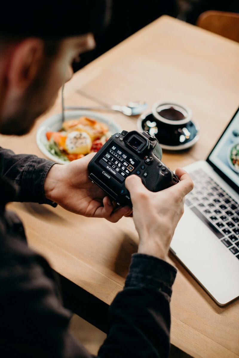 A man wearing black long sleeves is changing the settings on a black DSLR camera near a brown wooden table