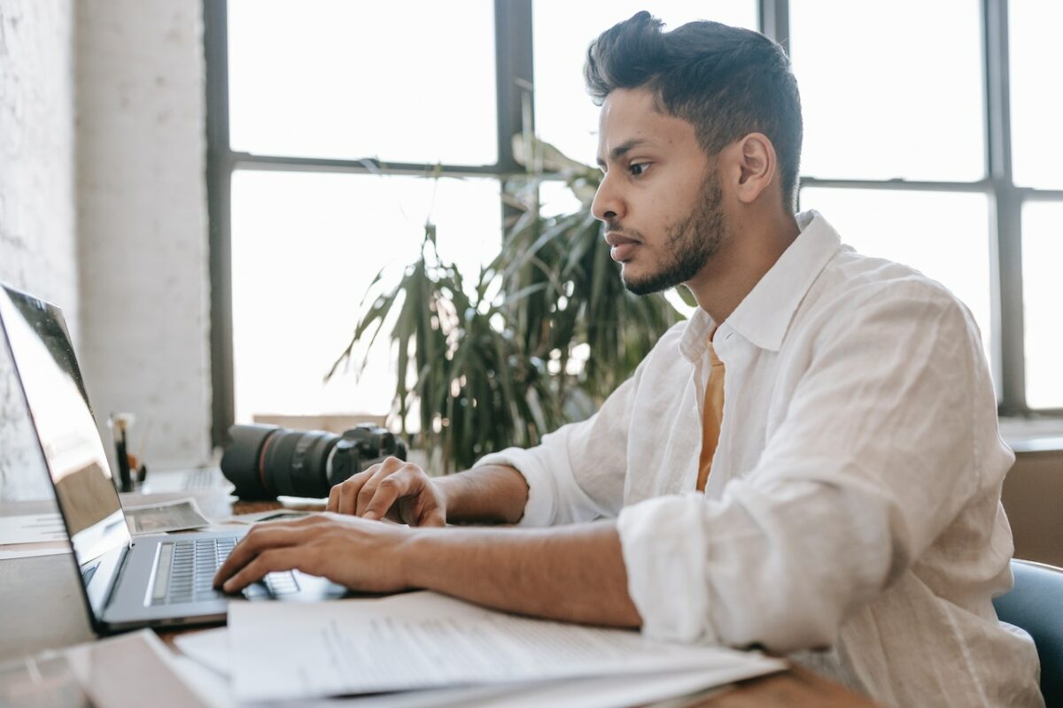 A man in white long sleeves working on a black laptop beside a black DSLR camera