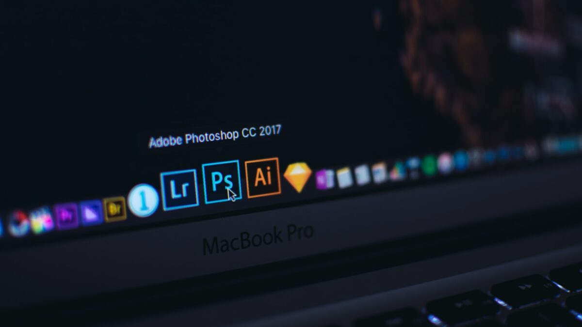 Un MacBook Pro argentato con sfondo nero e puntatore del mouse nero aleggia sopra il logo blu di Adobe Photoshop