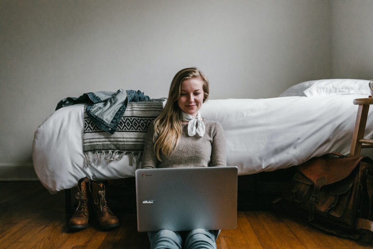 A woman wearing a brown long sleeves shirt and white scarf is seated on a brown wooden floor while using a silver Acer laptop