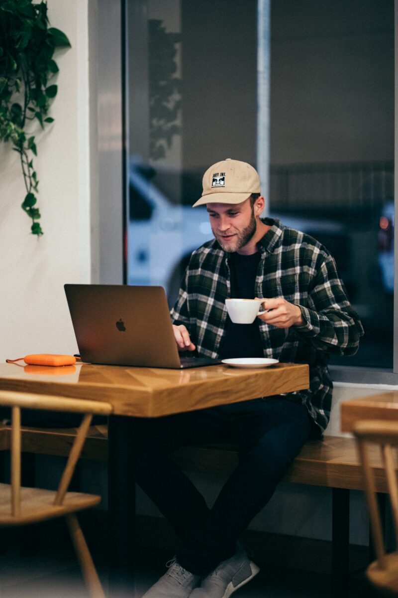 A man wearing a black and white plaid shirt is using a MacBook on top of a brown wooden table while holding a white mug