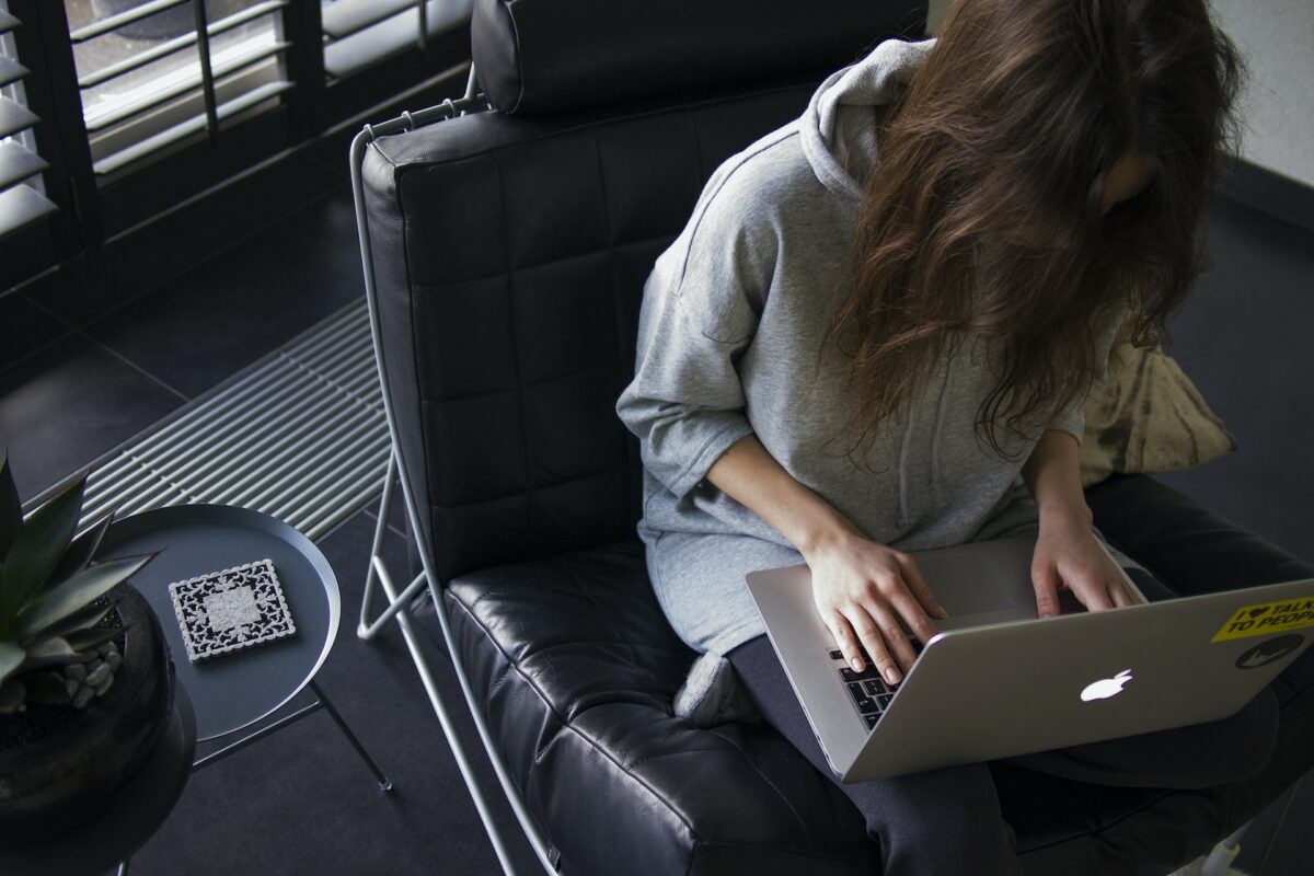 A woman wearing a gray hoodie is using a silver MacBook while sitting on a black leather chair