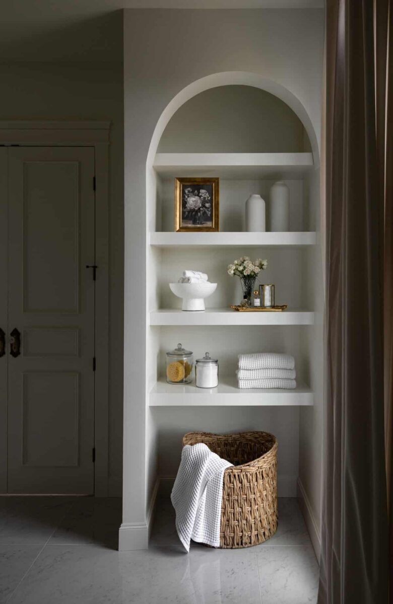 A white arched shelving with brown wooden laundry basket and white towels near a white door.