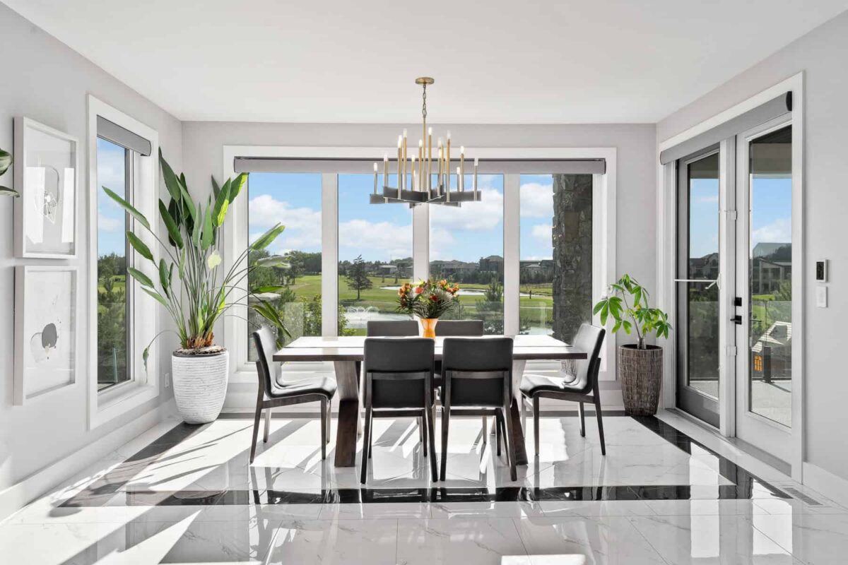 A bright dining area with large windows overlooking a green landscape, featuring a table, chairs, and decorative plants.