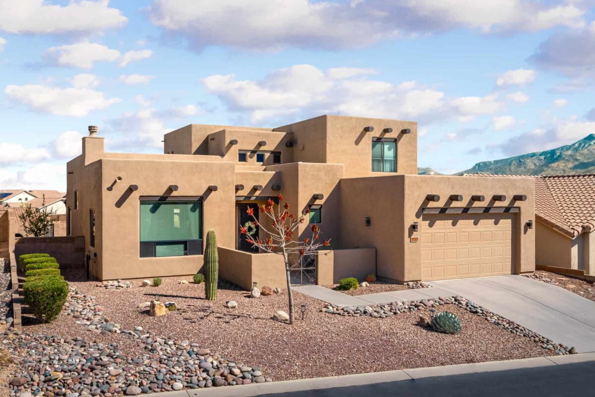 A modern southwestern-style house with beige stucco, desert landscaping, and a garage, set against a mountain backdrop.